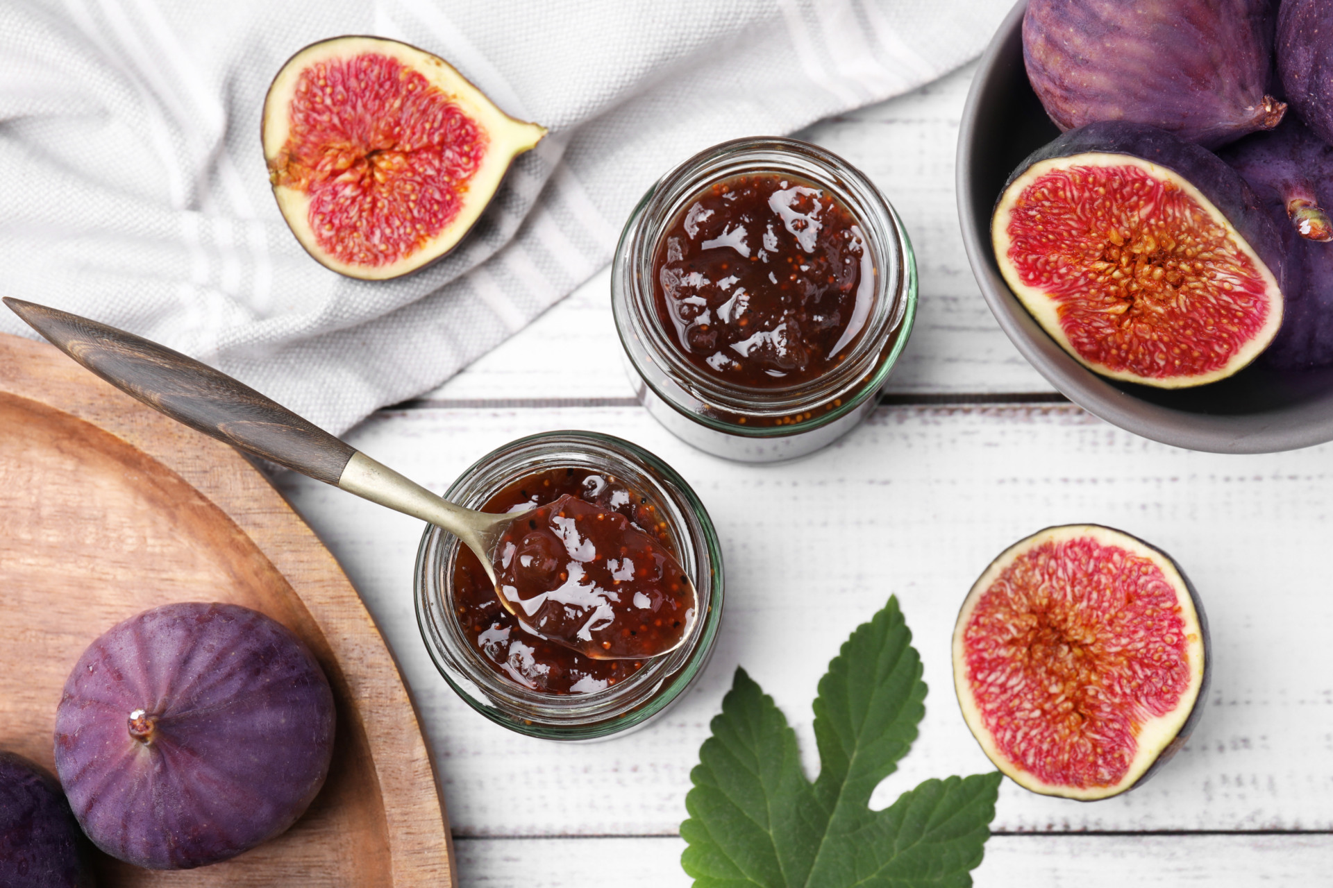 Glass jars of tasty sweet fig jam and fruits on white wooden tab